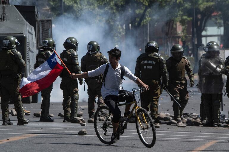 A protester on a bicycle waves a Chilean flag during clash with riot police in Santiago, on October 21, 2019. - Chile ordered an overnight curfew for the third day in a row on Monday as violent demonstrations and looting that left 11 people dead continued for a fourth straight day. (Photo by Pedro UGARTE / AFP)