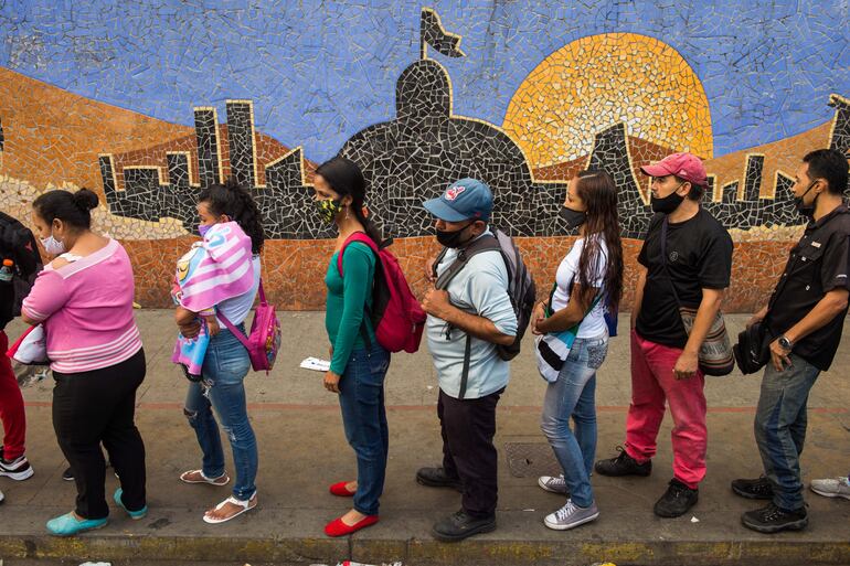 Personas en fila para abordar un bus frente a un mural que muestra la sede de la Asamblea Nacional de Venezuela, en Caracas.