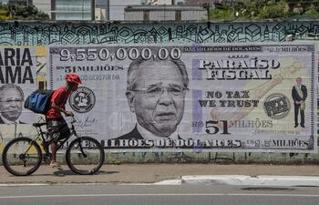 A man rides his bike past a poster depicting Brazil's Economy Minister Paulo Guedes on a US$9.55 million bill, in Sao Paulo, Brazil, on October 8, 2021, just days after he was mentioned in the "Pandora Papers" media investigation exposing world leaders' use of tax havens. - According to the investigation, Guedes has deposited US$9.55 million in his offshore Dreadnoughts International Group, opened in 2014 in the British Virgin Islands, which was equivalent to R$23 million at the time. With the devaluation of the real, accelerated during his tenure at the Ministry of Economy, the value today corresponds to R$51 million. (Photo by NELSON ALMEIDA / AFP)