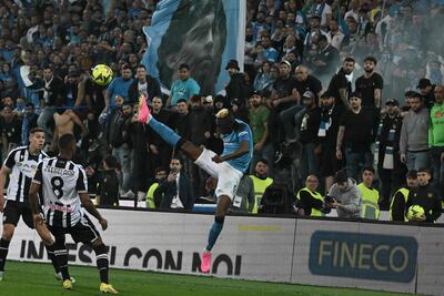 Víctor Osimhen puntea el balón ante dos jugadores rivales y con una imagen de Diego Maradona en el fondo, en las tribunas del estadio de Udinese, durante el partido de ayer que coronó campeón al Napoli.