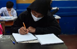 Un alumno con tapabocas completa su cuaderno en el salón de clases.