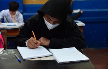 Un alumno con tapabocas completa su cuaderno en el salón de clases.