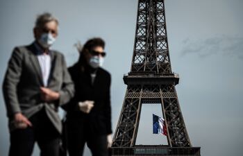 Un hombre y una mujer con máscaras faciales caminan en la Plaza del Trocadero mientras una bandera nacional francesa ondea en la Torre Eiffel.