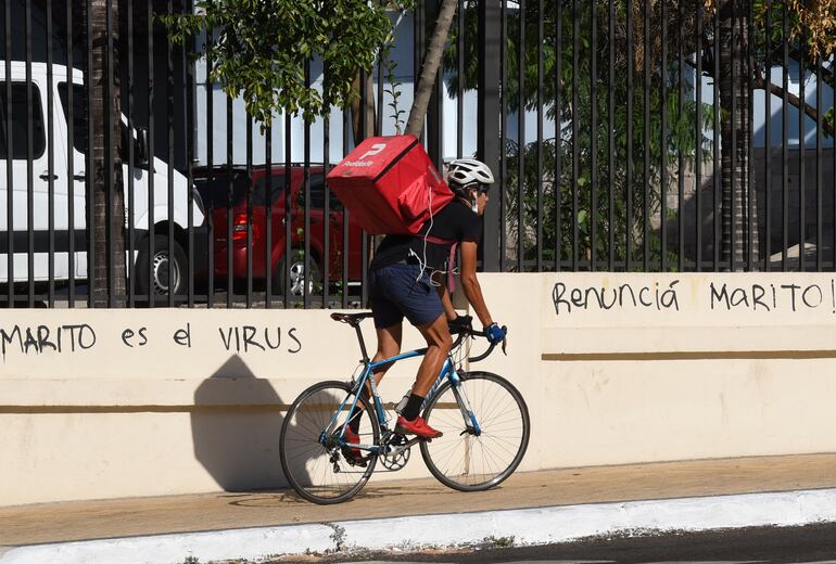 Un repartidor de comida circula en bicicleta por la vereda de la avenida Mariscal López, cerca de Mburuvicha Roga. En la pared se lee "Marito es el virus" y "Renunciá Marito". Al quedar desempleadas, muchas personas comenzaron a trabajar en el servicio de entregas puerta a puerta.