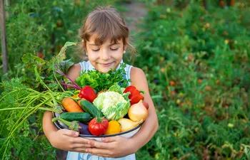 Una niña de unos 8 años, parada en medio de una huerta verde, sonríe mientras sostiene un bowl cargado con verduras como zanahorias, lechuga, coliflor, pepino, cebolla, tomate, berenjena y más.
