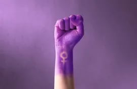 Un puño en alto de un brazo de mujer, teñido de lila, con el símbolo del feminismo.