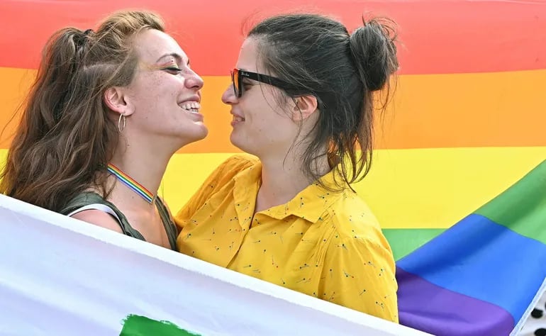 Dos mujeres se abrazan y propinan muestras de cariño frente a una bandera del orgullo gay.